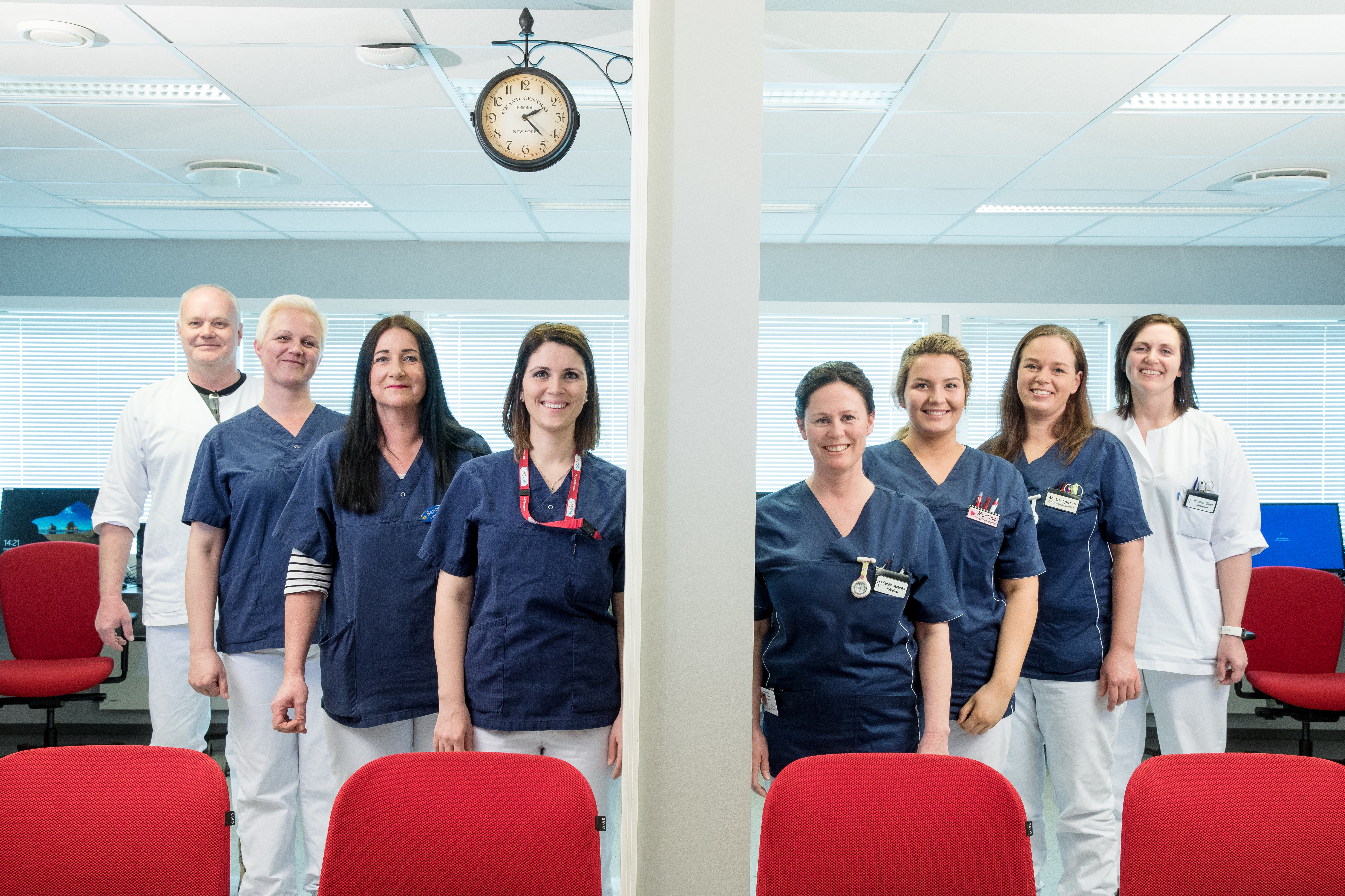 Gruppebilde smilende helsepersonell kledd i hvite og blå uniformer stilt opp bak røde stolrygger og klokke henger fra tak. Foto