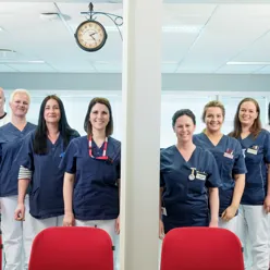 Gruppebilde smilende helsepersonell kledd i hvite og blå uniformer stilt opp bak røde stolrygger og klokke henger fra tak. Foto