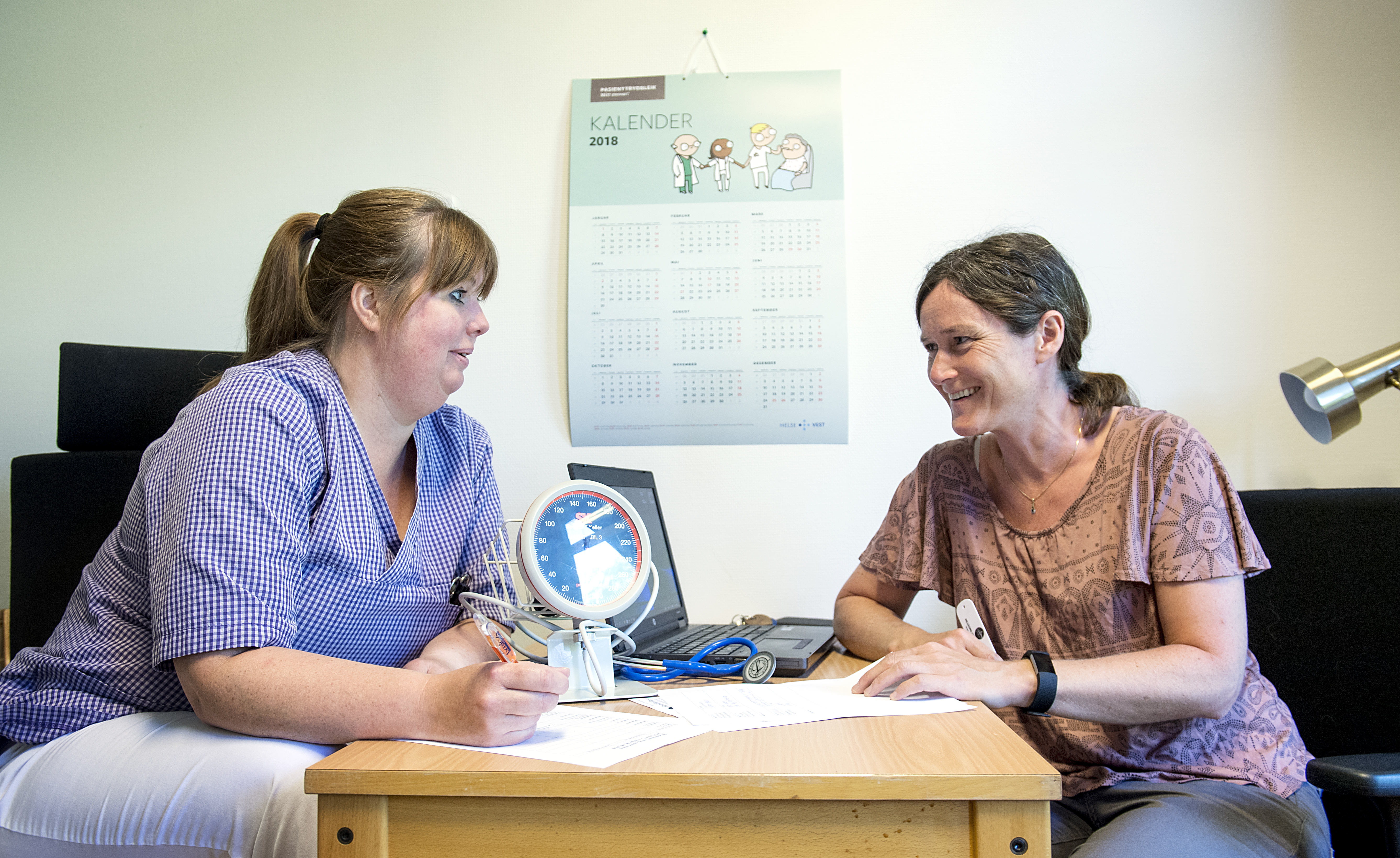 Foto: To personer prater sammen på legekontor med kalender på veggen.