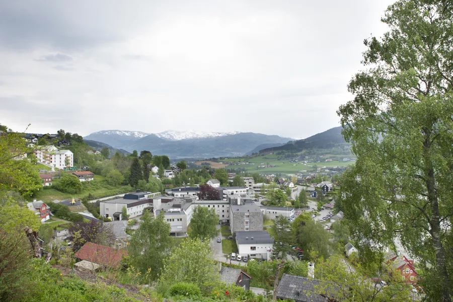 Oversiktbilde av sjukehusbygga til Voss sjukehus omgitt av grøne åsar og snøkledde fjell i bakgrunnen.